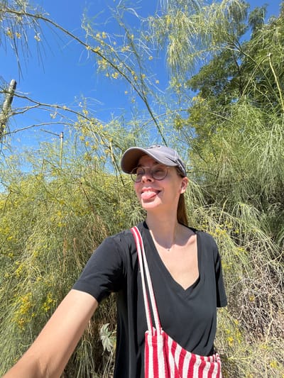 The image is a vertically oriented selfie taken outdoors. The main subject is a young person wearing a grey baseball cap, round glasses, and a black shirt with a red and white striped trim. They are sticking their tongue out and smiling, looking towards the camera. A thin necklace is visible around their neck.
The setting appears to be a dry, arid landscape with abundant green foliage and small yellow flowers, possibly a desert or semi-arid region. The sky is a clear, bright blue, suggesting it is daytime with sunny weather. The vegetation is dense and appears to be thorny or spiky. The orientation of the photo implies the person may be lying down or is positioned at an unusual angle relative to the typical upright perspective. The overall mood is playful and casual, indicative of a relaxed outdoor activity like hiking or exploring.
No visible text is present in the image. The location context provided is Azor, Israel, which aligns with the visual cues of a sunny, arid environment.