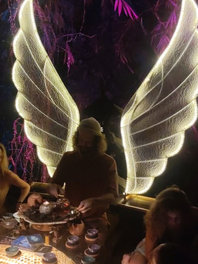 In a dimly lit outdoor setting at Lokaserena, Indonesia, a group of individuals gathers around a low table. A bearded man wearing a beanie is positioned centrally, his hands engaged in preparing or serving tea from a collection of teapots and cups arranged on a tiered platter. The table is covered with a patterned cloth. To his left, a woman with blond hair has part of her arm showing in frame. To his right, a dark-haired woman with curly hair sits facing down. Behind the central figure are large, illuminated angel wings, adding to the ambience of the scene. The background features dark foliage with sparse, purplish leaves, possibly bamboo.