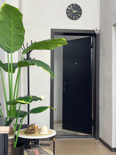In an indoor entryway, a large artificial plant with broad green leaves, tinged with yellow along the edges, dominates the left side of the frame. To the right, a dark grey doorway is partially open, revealing a glimpse of a hallway beyond. A black door with a small circular peephole and a silver handle is visible within the doorway. Above the doorway, a circular black clock with gold numbers and hands hangs on a light grey wall, displaying the time as approximately 9:42. A small decorative item, resembling a frog perched on coins, and a white candle with writing are placed on a small white table near the plant. A coat rack stands behind the plant. The flooring appears to be tiled. The overall scene depicts a domestic interior with decorative elements.