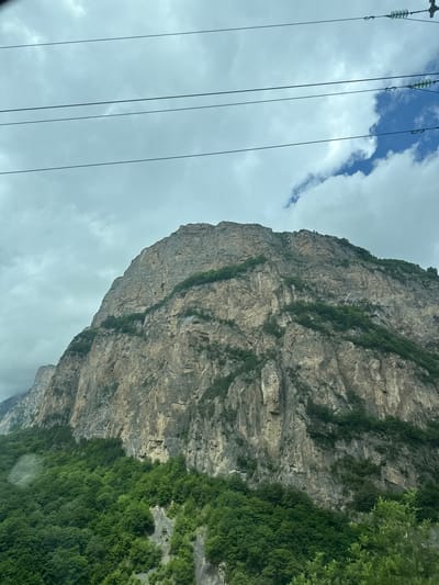 This image captures a dramatic mountain landscape, likely taken from a moving vehicle given the perspective and the presence of power lines in the foreground. The dominant subject is a sheer, massive rock face, its surface weathered and textured with visible strata and patches of greenery clinging to its crevices. To the right, dense green foliage, possibly a forest, cascades down the lower slopes of the mountain.

In the upper left portion of the image, a cloudy sky is visible, with breaks of blue sky and wispy clouds. The power lines, strung with glass insulators, cut diagonally across the frame, suggesting a developed area or route near the natural landscape.

The scene is likely set during the daytime, given the ambient light illuminating the mountain. The cloudy weather creates a slightly diffused light, casting soft shadows on the rock face. The raw, imposing nature of the mountain and the presence of man-made power lines hint at a location where human infrastructure interfaces with rugged natural environments. There are no people or discernible activity taking place within the frame. No text is visible in the image. The overall impression is one of natural grandeur and the contrast between the raw power of the mountain and the human presence implied by the power lines.