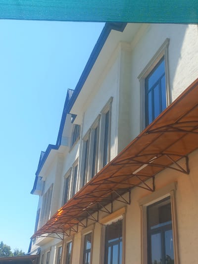 The image captures an architectural detail of a building with a clear blue sky as the backdrop. The building features several windows, some of which appear to be skylights, reflecting the bright sky. An orange awning, supported by a metal frame, extends over part of the building, casting a shadow. The architectural style suggests a modern design with clean lines and a light-colored facade.
A turquoise mesh screen is visible on the left side of the image, partially obscuring the building and sky. The overall scene is bathed in bright sunlight, indicating it is daytime, likely late morning or early afternoon given the intensity of the light and the shadows. The weather appears to be clear and sunny.
The image is taken from a low angle, looking upwards at the building. There is no activity or people visible in the frame. The location context of Yorkatay, Uzbekistan, combined with the architecture, might suggest a building in a region with a hot climate, hence the awning for shade. No visible text is present in the image.