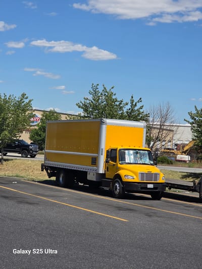 An eye-level, vertical, medium shot captures a bright yellow box truck parked on asphalt with yellow lane markings. The truck is positioned to the right of the frame, facing towards the viewer's left. The truck's cargo area is a large, bright yellow box with silver trim along the top and vertical supports. The truck cab is also yellow with a silver grille and black bumper. The windshield is covered with a reflective sunshade. A license plate with the inscription "EP17599" is visible on the front bumper. To the left of the truck, a black pickup truck is partially visible, driving away from the camera. In the background, a building with the word "UTILITY" in a circular logo is visible, surrounded by trees. Further in the background, construction equipment, including a yellow excavator, is present. The sky is blue with scattered white clouds. The image has a watermark in the bottom left corner that reads "Galaxy S25 Ultra." The scene appears to be an outdoor commercial or industrial area.