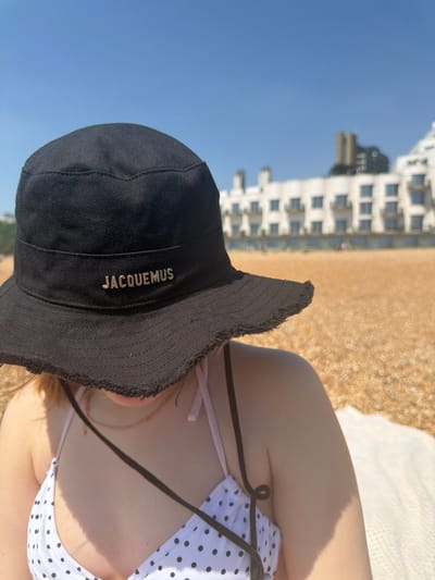 A daytime eye-level shot captures a person wearing a black "JACQUEMUS" branded hat and a white and black polka dot bikini top. The person is on a beach with light brown sand and a white object behind them, possibly a towel. In the background, a multi-story white building with multiple windows is visible, suggesting an urban beach scene in Folkestone and Hythe District, United Kingdom. The sky is clear and blue.