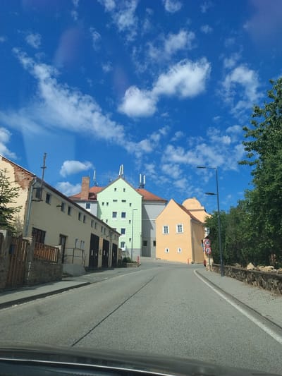 The image depicts a street scene in Lhenice, Czechia, under a bright blue sky with scattered white clouds. The perspective is from a vehicle driving down a paved road. On the left side of the road, there is a row of buildings with yellow and light green facades. The yellow buildings have red-tiled roofs and feature multiple garage doors. One of these buildings shows exposed brickwork on its upper portion. A wooden fence and stone wall line the property in front of these buildings. Further down the street, a light green building stands prominently with a red-tiled roof and numerous windows. On the right side of the road, a row of trees with dense green foliage can be seen. A street lamp with two lights is positioned on the sidewalk. Two circular traffic signs, one indicating a speed limit of "30" and another showing a prohibitory symbol with an arrow, are mounted on a pole near the sidewalk. The road curves gently to the right in the distance. The overall scene suggests a quiet, residential or commercial area in a European town.