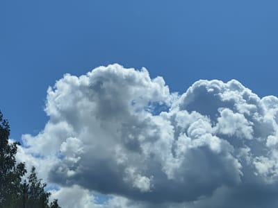 A bright blue sky is visible in the background, with a few fluffy white clouds scattered across the upper portion. The bottom of the image features a dark green tree on the left side and large, dense clouds that appear to be either cumulus or cumulonimbus. These clouds are a mix of white and gray, suggesting they may be on the verge of producing rain. The overall scene depicts a partly cloudy day in Lielvārži, Latvia.