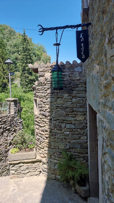 This image depicts a rustic stone building with a hanging sign and lantern. The sign, a black banner with white lettering, reads "La teca". A green glass lantern hangs from a wrought-iron fixture, which also supports the sign. The building is constructed of rough-hewn stones, characteristic of the medieval architecture found in Rupit i Pruit, Spain, a village renowned for its well-preserved Catalan Romanesque and Gothic buildings. The background features lush green trees and a clear blue sky. A lamppost stands to the left, and a stone planter with greenery is visible in the foreground. The scene suggests a historical setting, likely a shop or restaurant, given the signage.