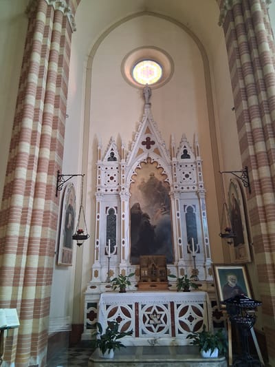 This is a photograph of the interior of a church in Padua, Italy, specifically the altar area. The focal point is a highly ornate white altar with decorative carvings and a large painting above it depicting religious figures. To the left and right of the altar are tall, striped columns, alternating between light and dark colors, possibly brick and stone.

Above the altar painting, a circular stained-glass window is visible in the pale wall, illuminated by natural light. A small statue of a robed figure stands on a decorative pinnacle above the painting. Hanging from iron brackets on the walls to the left and right of the altar are ornate lanterns with what appears to be red candles inside.

On the left wall, a framed painting of a robed figure is hanging. On the right side of the altar, a framed portrait of an older man with a beard, wearing religious attire, is propped up on an easel. In front of this portrait is a dark metal stand with several small candle holders.

The altar itself is covered with a white lace cloth. The floor in front of the altar is tiled in a black and white checkered pattern. Potted green plants are placed on the steps leading up to the altar and on the altar itself. A lectern with an open book is visible on the far left of the image, near the striped column. The overall impression is one of solemnity and religious devotion.
