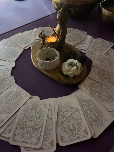 A close-up shot displays a collection of tarot cards arranged in a circular formation on a textured, purple surface. At the center of the circle lies a wooden board holding a white, floral candle, a small candle burning beside a Buddha statue, incense, and a white, ridged container. The overall setting suggests a spiritual or ritualistic atmosphere. It is not possible to determine the location of Solovej-Kljuch, Russia from this image.
