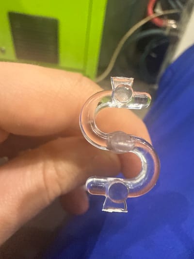 A person's hand is holding a clear, plastic "S" shaped object. The object has small circular protrusions at each end and in the middle. The background includes a green surface, some wires and a blue surface. The location is not discernible from the image.