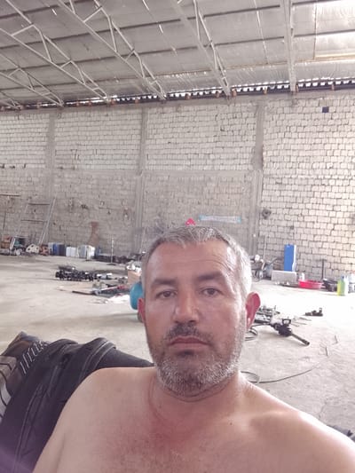 The image captures a man taking a selfie indoors in what appears to be a workshop or industrial building. The man, with graying short hair and a gray beard, looks directly at the camera with a neutral expression. He is depicted shirtless, showing his chest and shoulders, and is seated in a dark, possibly leather, chair or car seat.
The background reveals a spacious room with light-colored brick or cinder block walls, showing some signs of wear and concrete support beams. Above, a high ceiling features an exposed metal truss structure supporting what appears to be a corrugated metal roof, possibly with reflective insulation panels. The concrete floor is cluttered with various objects, indicating a work environment. These include several machinery parts (such as crankshafts), a ladder leaning against a wall, plastic containers (blue, white, red), and an impact wrench with a power cord.
The setting suggests a functional garage, repair shop, or a similar industrial facility, characterized by its unfinished aesthetic and scattered tools/parts. The ambient lighting indicates it is daytime. Faint, unreadable text can be observed on one of the brick walls in the background. The provided location context for this scene is Sabir Rakhimov, Uzbekistan.