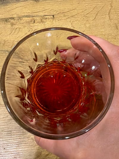 A close-up, top-down view shows a person's hand holding a faceted glass containing a red liquid. The glass has decorative etchings on its sides. The background is a wooden surface with a visible grain. The scene is indoors, not in Tabasalu alevik, Estonia.