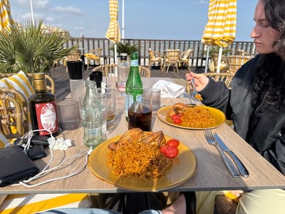 The image depicts an outdoor dining scene on a wooden deck in Los Alcázares, Spain, under a clear, bright sky. The setting features yellow and white striped umbrellas and cushions, wicker chairs, and visible palm foliage, with a railing overlooking water in the background.
On a light wood-grain table, two identical meals are served: each plate contains a substantial portion of an orange-colored rice dish, likely paella, alongside a roasted chicken quarter and three halved red cherry tomatoes. A person with dark, curly hair, wearing a black garment, is partially visible on the right, actively eating from one of the plates with a fork raised to their mouth.
The table also holds an "Armonia Aceite de Oliva" bottle, an empty clear Coca-Cola glass bottle, a green glass bottle (possibly sparkling water), several glasses (one containing a dark beverage, others empty), a black salt or pepper shaker, and a crumpled white napkin. Personal items including a black power bank, white charging cables, and keys are positioned on the left side of the table. A fork and knife set are neatly placed next to one of the plates.