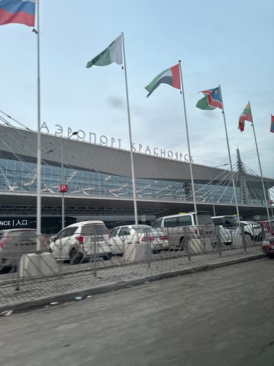 The image shows the exterior of the Yemelyanovo International Airport in Krasnoyarsk, Russia. The modern airport building is visible, with a facade of glass and metal. The Cyrillic script "АЭРОПОРТ КРАСНОЯРСК" (Airport Krasnoyarsk) is displayed prominently on the building. Several flagpoles are lined up in front of the building, each displaying a different flag. Cars are parked along the curb, separated from the sidewalk by a metal barrier. The sky is overcast. The image is slightly rotated.