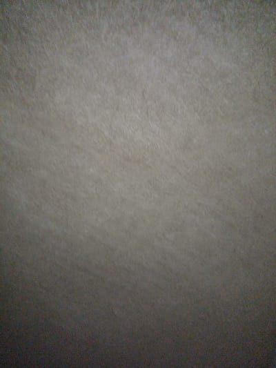 The image is a close-up, slightly blurry shot of a textured surface. The surface appears to be a light brown or beige material with subtle variations in tone and visible fiber-like patterns. There is a gradient of light, with the top portion of the image being brighter and gradually becoming darker towards the bottom. The texture suggests it could be paper, fabric, or a natural material. There are no people, objects, or any discernable environmental features visible in the image. The image provides no information that would allow for the identification of a specific location within the city of Veremia, Ukraine.