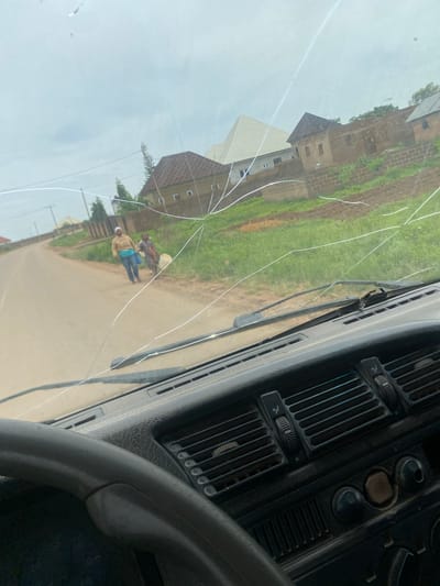 The image shows a view from inside a vehicle, looking through a cracked windshield at an outdoor scene. The dashboard of the vehicle is visible in the foreground, with parts of the steering wheel and air vents. Outside, a paved road curves to the left, bordered by a grassy verge. Two individuals, appearing to be women, are walking along the road. One is wearing a tan jacket and blue pants, and the other is wearing a patterned dress. Behind them, there are several buildings with brick walls and varied roofing styles. The sky is overcast. The location is identified as DU, Nigeria.