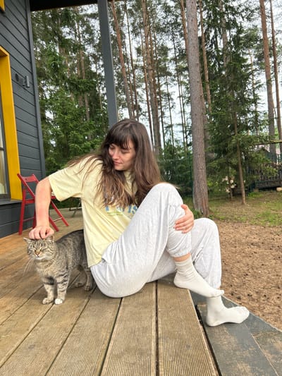 A young woman with long brown hair, wearing a light yellow t-shirt and light grey sweatpants with white ribbed socks, is sitting on a wooden porch. She is petting a tabby cat that is standing next to her. The woman is looking down at the cat, who has its eyes closed and appears to be enjoying the attention. The porch is made of wooden planks and is surrounded by tall pine trees. The house to the left is dark grey with a bright yellow window frame. In the background, there is a fence and a pile of firewood. The location is the city of Shepelyovo, Russia. The overall scene is peaceful and relaxed.
