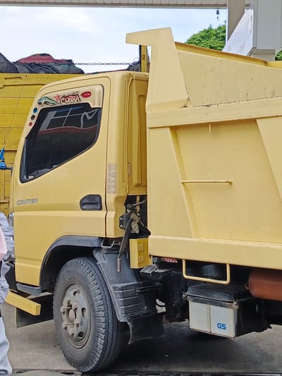 The image shows a yellow dump truck parked outdoors. The truck has "CANTER" written on the side of the cabin, and a stylized "COBRA" logo above the driver's side window. The truck's bed is large and yellow. On the right side of the truck, near the ground, a battery pack with "GS" branding is visible. The truck appears to be in an urban environment, possibly a roadside or parking area, given the concrete ground and the suggestion of a canopy or structure overhead. No specific landmarks or details clearly identify the location as Cotbadatunong, Indonesia.