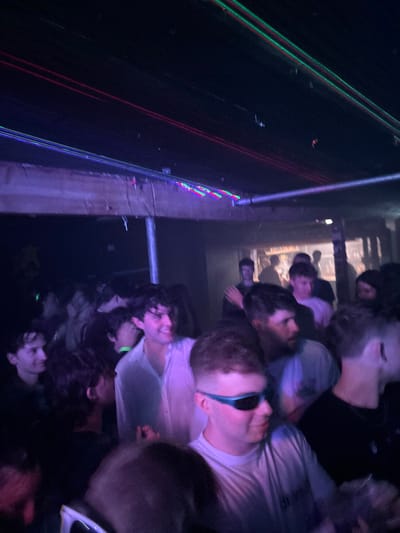 A blurry, angled shot captures a dimly lit event with numerous individuals, likely a party or concert. Laser beams, colored red, green, and blue, streak across the dark space. People cluster close together, their faces partially visible. Illumination from above casts a purple hue across the scene. A metal railing is visible in the frame as well.