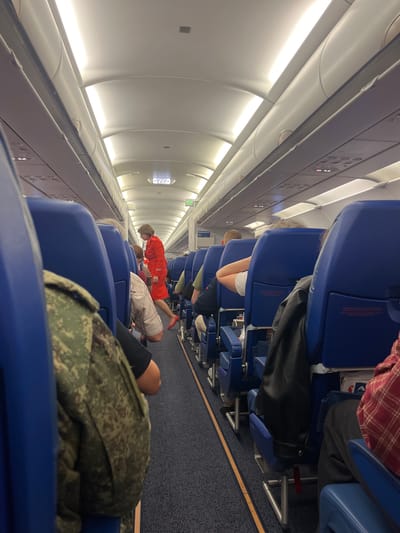 The image shows the interior of an airplane cabin, viewed from the perspective of a passenger. The seats are blue, and the aisle is carpeted in dark blue with a yellow stripe. Overhead compartments are visible, along with interior lighting strips. A flight attendant in a red uniform is walking down the aisle, facing away from the camera. Several passengers are seated, their backs to the camera, some wearing casual clothing, and one person in a camouflage jacket is in the foreground on the left. An "EXIT" sign with Cyrillic lettering is illuminated above the aisle further down the cabin. The scene depicts a typical commercial flight environment.