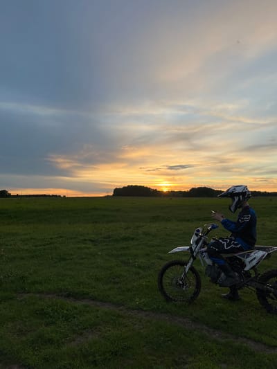 The image shows a person on a motorcycle in a grassy field, with a sunset in the background. The person is wearing a helmet and riding gear. The motorcycle is white with black and blue accents. The grass appears to be in a field with some paths, possibly in the region of Новочувашский, Russia. The sky is partially cloudy, with colors of orange, yellow, and blue indicating a sunset.