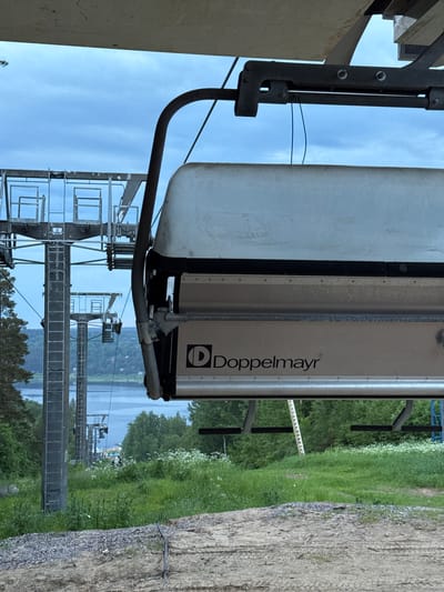 An unoccupied Doppelmayr chairlift seat, silver with black trim, hangs suspended from a dark metallic support arm in the foreground. Two blue wires hang loosely from the chairlift mechanism. Above, a light-colored, flat overhead structure is partially visible. The background reveals an overcast light blue sky above a large body of water, flanked by a densely forested distant shore. Multiple steel lattice chairlift towers descend a green, tree-lined slope towards the water. The closest tower is marked with the letter "B". The immediate foreground consists of a grassy area interspersed with white wildflowers, leading to a section of disturbed earth and gravel. The scene depicts a ski resort in Korobitsyno, Russia, during a non-operational period. No people are present.