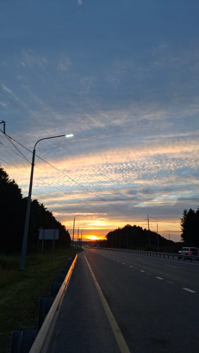 The image captures a scenic view of a highway in Bogandinsky, Russia, during sunset. The sky is painted with a gradient of warm colors, ranging from vibrant orange and yellow near the horizon to soft pinks and blues higher up, interspersed with wispy clouds. The sun is setting on the left side of the frame, casting a warm glow on the surroundings.
The highway itself is dark asphalt with a prominent yellow line marking the center. A silver guardrail runs along the right side of the road, reflecting the sunset's light. Trees form a dark silhouette along the left edge of the highway, bordering the sunset sky. Several streetlights are visible, with one in the foreground on the left displaying a bright white light. In the distance, on the highway, a white vehicle can be seen.
The overall impression is one of tranquility and the beauty of nature at the end of the day. The setting is rural or semi-rural, suggested by the presence of trees and the open highway. The time of day is clearly sunset due to the lighting and the colors in the sky. There is no visible text in the image.