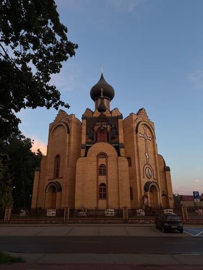 The image depicts the exterior of an Eastern Orthodox church, constructed primarily of yellow brick, situated in Sacharewo, Poland. The church features a large, dark, onion-shaped dome topped with a cross, and smaller domes or architectural elements. The facade is adorned with a prominent cross and circular icons. Windows are arched, and wooden doors are visible. A metal fence with brick pillars separates the church grounds from a paved area. Trees, including one with dark foliage in the foreground, are present to the left and behind the structure. A dark-colored hatchback car is parked in a designated parking space in front of the church. The sky is a gradient of blue and pink, suggesting either dawn or dusk. A street sign indicating parking and accessibility is visible to the right of the car. The scene is devoid of any people.