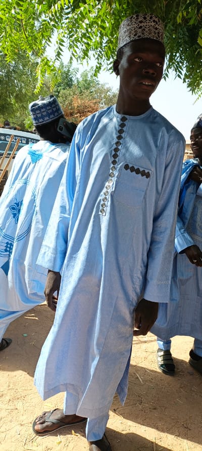 The image depicts an outdoor scene in Ungongo, Nigeria, under bright daylight. A young Black male stands centrally in the foreground, facing slightly right of the camera with a neutral expression. He wears a light blue, long-sleeved traditional garment (likely a kaftan) with a subtle diamond pattern woven into the fabric. The garment features a vertical line of 14 dark gray circular decorative buttons or studs down the front, from the neck to the mid-chest. A breast pocket on the left side (viewer's right) is adorned with five similar circular dark gray embellishments. He wears a white kufi cap with an intricate dark brown or black pattern on his head. His feet are clad in brown flip-flops.
To the left and slightly behind the main subject, another male figure is partially visible from the back, wearing a light blue traditional garment with prominent darker blue embroidered designs. He wears a blue and white patterned kufi cap and is holding a dark-colored smartphone to his right ear, appearing to be in conversation. Further in the background, a light-colored vehicle, possibly a bus, is partially visible.
To the right of the main subject, a third individual is partially visible, also in a light blue garment, with a partial smiling face and a ring on their right hand. Their feet show black shoes with three straps. The ground is composed of dry, reddish-brown dirt with scattered light debris. Above, a canopy of green leaves from a tree provides partial shade, with a clear blue sky visible in the upper background.