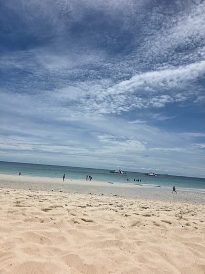 This image depicts a serene beach scene in Bang Saray, Thailand, under a dynamic sky filled with wispy white clouds against a clear blue backdrop. The foreground is dominated by textured sand, showing subtle ripples and indentations, suggesting a gentle breeze. In the mid-ground, a shallow, turquoise ocean gently laps at the sandy shore. Several people are visible enjoying the water and the beach. In the distance, two catamaran-style boats, each with attached water slides, are moored in the clear water, indicating a popular spot for recreational activities. A lone figure is seen jogging along the shoreline on the right side of the frame, while other individuals are scattered further down the beach, some wading in the shallows. The overall impression is one of a tranquil and inviting coastal environment.