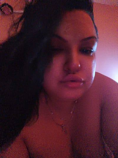 The image is a close-up, slightly blurry selfie of a person with dark hair, taken indoors. The lighting appears to be artificial, casting a warm, reddish-pink hue over the scene. The person's face is partially visible, with one eye closed or squinting. They are wearing a necklace with a pendant. The background is indistinct, but appears to be a wall or fabric. There are no discernible landmarks or architectural features that would indicate a specific location within the city of Komotini, Greece.
