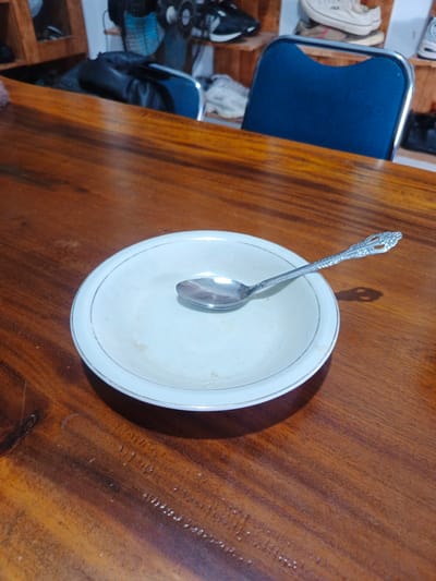 The image displays a white, shallow ceramic bowl with a gold rim, containing a silver spoon, centered on a polished wooden table. The bowl shows faint light brown stains or residue on its surface, indicating prior use. The spoon has a decorative, ornate handle design.

The wooden table features a rich, reddish-brown finish with visible grain patterns and some minor imperfections. The background is softly blurred, revealing an indoor setting. To the right of the table, the back of a blue chair with a metallic frame is visible. Further in the background, several pairs of shoes, including sneakers, are arranged on wooden shelves or racks, suggesting a storage or display area.

No specific activity is occurring, and no people are present in the frame. The lighting suggests an indoor environment. There is no visible text. The scene is located in Bireuenmeunasahcapa, Indonesia.