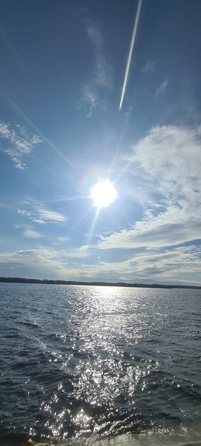The image presents a bright outdoor landscape, vertically oriented but likely intended as a horizontal view, showcasing a sky on the left and a body of water on the right, separated by a distant horizon. The setting is consistent with a natural environment near Ikšķile, Latvia, perhaps along the Daugava River.

The left portion of the image is dominated by a clear blue sky, interspersed with scattered white clouds that range from light, wispy formations to more textured, elongated patches. A brilliant, intense sun is centrally positioned in the upper-middle left of the sky, radiating strong light rays and creating a prominent lens flare with colorful, rainbow-like refractions. A distinct, straight white contrail extends diagonally across the upper left quadrant, marking the path of an overhead aircraft.

The right side of the photograph features a vast body of water, its surface glistening intensely with countless sparkling reflections from the sun. These shimmering highlights suggest active ripples or small waves on the water. A thin, dark line of land or distant shoreline is visible along the horizon line that horizontally bisects the image, creating a subtle boundary between the sky and water in the distance. This distant land appears to be covered in trees or dense vegetation.

The activity captured is simply the natural interplay of sunlight, sky, and water. The strong sunlight and high position of the sun suggest it is mid-day, possibly morning or early afternoon. The weather is clear and sunny with some scattered clouds. No people or artificial objects are prominently featured aside from the distant contrail. There is no visible text in the image. The overall mood conveyed is one of bright, serene natural beauty.