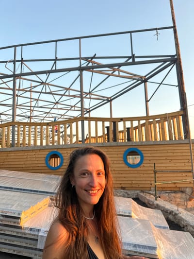 A young woman with long brown hair and fair skin is captured in a selfie taken outdoors. She has a wide smile, revealing her teeth, and her eyes are looking directly at the camera. She is wearing a dark-colored top and a thin necklace. The background suggests a construction site. A large metal structure, likely a framework for a building, dominates the left side of the image. To the right, a wooden wall with horizontal slats and two circular blue openings is visible. In front of the wooden wall and extending into the foreground are stacks of large, flat objects wrapped in clear plastic sheeting. Text is visible on the plastic wrap, including "ROCKPUR CE" and "SANDWICH PANELS". The sky is clear and blue, indicating daytime with fair weather. The overall scene appears to be in a rural or semi-rural setting, consistent with the location context of Svinjišta, Montenegro, and the activity of construction. The woman's expression conveys happiness and a sense of pride or engagement with the ongoing project.