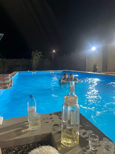 This nighttime image captures an outdoor scene in Yeghvard, Armenia, featuring a rectangular swimming pool with illuminated blue water. Two adult males are partially submerged in the pool, facing the viewer. The male closer to the left side of the frame has dark hair and a beard, with his right hand extended in a gesture. The male to his right, positioned slightly further back, also has dark hair and a neutral expression.
The pool deck, constructed of a light-colored material, occupies the foreground. On it, a tall, clear glass decanter approximately half-filled with a yellowish liquid stands prominently. Adjacent to it are a clear plastic water bottle and a ribbed glass tumbler containing a small volume of liquid. Two smaller clear shot glasses, one containing liquid, are positioned near the decanter. A hand holding a glass with a clear liquid extends into the upper left quadrant of the frame. A white, textured, low-lying object is partially visible in the bottom center.
The background consists of a dark night sky. A street light is visible mid-left, positioned beyond a dark, low wall and sparse trees. A bright artificial light source illuminates the pool area from the right, revealing a light-colored building structure.