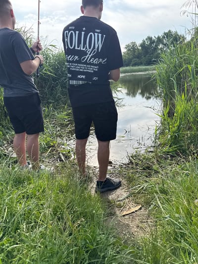 Two adult males are observed from the back, standing at the edge of a body of water in Zamfir, Bulgaria. The individual on the left wears a dark grey t-shirt, black shorts, and athletic shoes, actively holding a red and white fishing rod angled toward the water. A dark beaded bracelet is visible on his left wrist. The individual on the right wears a black t-shirt with prominent white text including "ESTD 1987 FOLLOW Your Heart" and motivational phrases, along with black shorts. He is positioned closer to the water's edge, appearing to be barefoot or wearing dark, low-profile footwear on muddy ground. The immediate environment consists of tall green grass and reeds, framing a narrow dirt path leading to the water. The body of water is calm, reflecting the partly cloudy sky and distant green trees, with some surface debris visible.