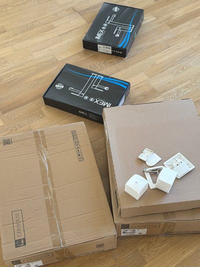 The image shows a collection of plumbing fixtures and their packaging on a wooden floor. Two black boxes, both labeled "IMEX BATH EQUIPMENTS," are positioned in the upper half of the frame, displaying line drawings of faucets. Below these, two large cardboard boxes are stacked. The top cardboard box has several small, white plastic components and some white, cube-shaped material scattered on its surface. The bottom cardboard box features branding for "CER HOUSE." The overall scene appears to be an indoor setting with a natural wood floor. There is no indication of the city of Gondomar, Portugal, or any specific location within it.