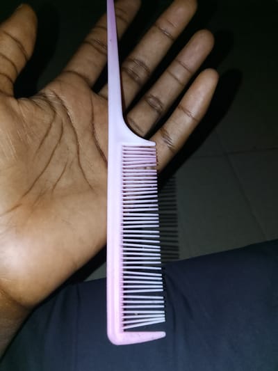 In Bida, Nigeria, a person's hand is holding a pink comb. The hand is open with fingers extended, and the comb is positioned vertically, with its teeth facing downwards. The comb is made of plastic and has a narrow handle. The background is dark and indistinct, suggesting an indoor or dimly lit environment.