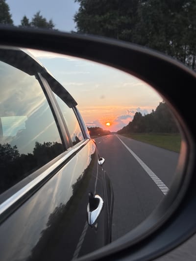 The image is a rearview mirror reflection showing a sunset over a highway. The reflection captures the sky with scattered clouds and the sun partially obscured by clouds, casting a warm orange glow. A dark-colored car is visible in the foreground, reflecting the sky and surrounding trees. The highway stretches into the distance, with clear lane markings. Dense trees line the right side of the highway. The overall scene suggests travel on a road in a rural or suburban area during twilight. There are no people visible in the image. The location is identified as Yassyenki, Belarus, which is a village in the Yassyenki rural council of the Mogilev Region in Belarus.