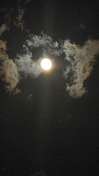 The image is a vertical, full-frame photograph of a bright, full moon partially obscured by clouds in a dark night sky. The moon is situated slightly above the center of the frame. Wispy clouds, illuminated by the moonlight, create a soft halo around the moon and extend across the upper portion of the image. The lower portion of the image is filled with the dark expanse of the night sky. There are no visible landmarks or indications of the city of Viterbo, Italy. The scene is devoid of any people, specific objects, or discernible actions.