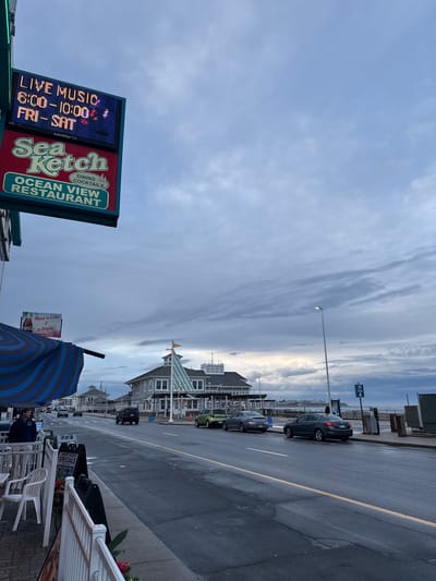 The image depicts a street scene in Hampton, United States of America. A sign for "Sea Sketch Ocean View Restaurant" advertises dining and cocktails with live music from 6:00-10:00 PM on Fridays and Saturdays. In the background, the street continues with cars parked alongside and buildings lining the road. A Coca-Cola sign is partially visible. The sky is overcast with varied cloud formations. An umbrella is present in the foreground. Street lights illuminate the scene. The photograph is taken at an angle, with the street and buildings slanting to the right.