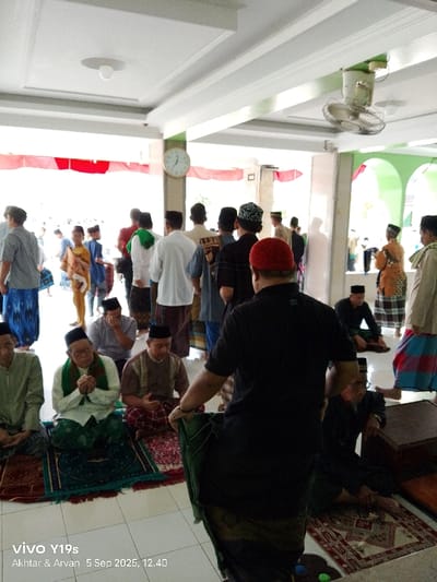 The image depicts an indoor gathering of predominantly male individuals within a large hall, identified as a mosque or prayer room, in Jagapura Kulon, Indonesia. The environment features a white tiled floor and a white ceiling with recessed lighting fixtures and exposed structural beams. A circular wall clock, showing approximately 12:40, is mounted on a pillar to the left. A white ceiling fan is visible in the upper right. Bright outdoor light enters from a large opening in the background, revealing green arched structures and red and white banners.
In the foreground, a man in a black polo shirt and a red skullcap stands with his back largely to the viewer, holding a green prayer mat. To his left, three men are seated cross-legged on individual prayer mats: one in a light green shirt, another in a white shirt with a green scarf and glasses, and a third in a brown patterned shirt. All three seated men wear black skullcaps, and the man in white has his hands clasped.
In the mid-ground and background, numerous other men and boys are standing or moving. Most are dressed in traditional Islamic attire, including sarongs (kain sarung), long shirts (baju koko), and various skullcaps (peci/kufi). They appear to be congregating, likely in preparation for or following a religious service.
