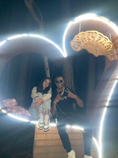 The image shows a low-angle shot of two people standing inside a lighted wooden structure in Sungurlare, Bulgaria. The structure is marked with "СУНГУРЛАРЕ," indicating the city name. One individual, a dark-skinned man, is positioned at the top of the frame, wearing sunglasses, a black T-shirt, and a chain necklace. He is making a "peace sign" with both hands. Below him, a light-skinned woman is sitting with her legs drawn up, wearing a light-colored long-sleeved shirt and denim shorts. A plaid blanket is draped over her legs. The structure appears to be a decorative arch or entrance, illuminated with bright lights around its edges, creating a tunnel effect in the photograph. The background is dark, suggesting an outdoor nighttime setting. The wooden planks of the structure are visible, and there are tree branches overhead.