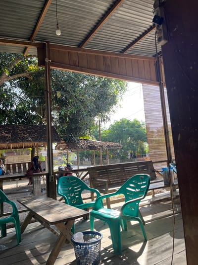 The image, when correctly oriented, depicts a simple, open-air structure, likely an eatery or waiting area, in Keudesampoiniet, Indonesia, during a bright, sunny day. The interior features a dark wooden plank ceiling from which a power strip with electrical sockets and wires is visible. On the left, a corrugated metal wall with wooden support beams holds a single hanging light bulb.

The floor on the right side of the structure is made of wooden planks, showing strong sunlight and sharp shadows. Several vibrant green plastic chairs are arranged around simple wooden tables. A blue plastic basket rests beside one of these tables.

Beyond the structure, the scene opens to a verdant outdoor area with lush green trees under a clear sky. A bamboo screen or blind hangs above the opening. In the mid-ground, a person wearing dark clothes and a head covering (hijab) is visible, standing or sitting on a lower platform, with a red motorcycle partially visible nearby. Another person is partially seen sitting towards the bottom right. The overall atmosphere is casual and relaxed, indicative of a typical daytime setting in a tropical region. No discernible text is visible.