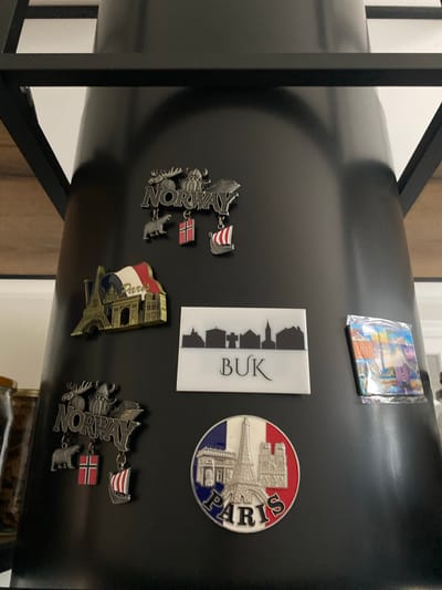 The image shows a collection of magnets attached to a black cylindrical object, likely a refrigerator door. The magnets represent souvenirs from different locations.

On the upper right, there is a circular magnet with a depiction of the Eiffel Tower and the word "PARIS" encircling the image. Next to it, to the left, is a rectangular white magnet with a silhouette of buildings and the word "BUK" printed vertically. Above "BUK" is a small, shiny packet of magnets, possibly a multi-pack souvenir.

Below the "BUK" magnet, to the left, are two magnets with Norwegian themes. One is a longer metal magnet with the word "NORWAY" and small metal figures of a moose and a bear. A small Norwegian flag and a Viking ship with a flag are attached by chains. To the right of this, slightly higher up, is a smaller Norwegian flag.

On the lower right, there is a metallic magnet featuring the Eiffel Tower with a small French flag and the word "Paris" written on it. To the right of the Eiffel Tower magnet, partially visible, is another magnet with Norwegian imagery, showing "NORWAY" with a moose, a bear, and a Norwegian flag.

The setting appears to be indoors, with a wooden floor visible in the background. The lighting suggests it's daytime. No people are visible in the image. The overall impression is a display of travel memories.