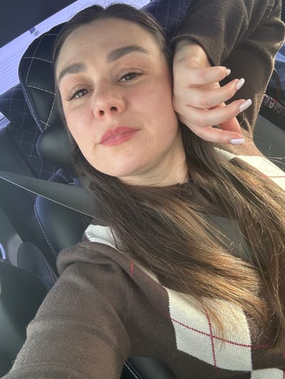 The image displays a single female individual inside the cabin of a vehicle. The specific location in the city of Mostovaya, Russia, cannot be determined from the visual information provided.
The subject has long, dark brown hair, centrally parted, cascading over her right shoulder and chest. Her complexion is light, with light pink lipstick on her lips and defined eyebrows. Her left hand, featuring manicured nails with an opaque, light pink or nude polish, is positioned against her left cheek and jawline. She wears a brown long-sleeved sweater adorned with a white and dark red argyle pattern across the chest and right arm. A black seatbelt is visible diagonally across her chest and left shoulder. The individual is looking directly towards the camera, suggesting a self-portrait.
The immediate background consists of a black car seat with a quilted pattern on the headrest, indicating a vehicle interior. A diffused light source, consistent with daylight, illuminates the scene from the right side, with a faint view of a rear window behind the subject's head.