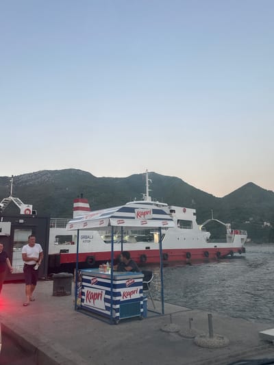 The image depicts a ferry docked at a pier in Jošice, Montenegro. The ferry, named "GRBALJ KOTOR," is white with a red hull and red and white striped smokestack. It is positioned alongside a concrete pier where several people are visible.

To the left of the ferry, a man in a white t-shirt and dark shorts is walking away from the camera, while a woman in dark clothing walks beside him. Further left, a portion of another ferry is visible.

In the foreground, a small kiosk with a striped blue and white awning is set up, selling drinks. The kiosk has a refrigerated display case branded with "Kapri." A man is seated behind the kiosk, and cans of "Frikon" are visible on display.

The background is dominated by a lush, green, mountainous landscape that slopes down to the water. The sky is clear and pale blue, suggesting it is either early morning or late evening. The water is calm. The overall scene is of a coastal town with active ferry transportation.