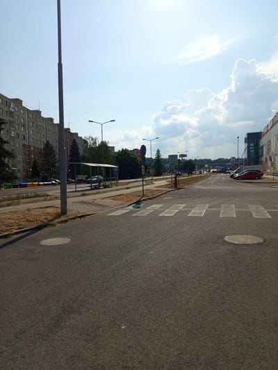 The image shows a street intersection in Zvolen, Slovakia, on a sunny day. In the foreground, there is a paved road with a pedestrian crossing marked by white stripes. Two circular manhole covers are visible on the road. To the left of the road, there is a sidewalk with dry grass and a lamppost. Further back, a bus stop with a shelter and a bench is situated. Beyond the bus stop, there is a row of apartment buildings and trees. In the background, more buildings, including a large commercial building with a "BILLA" supermarket logo, are visible. Several cars are parked and driving on the road. The sky is blue with some white clouds. The scene appears to be a typical urban environment with elements of public transport, residential areas, and commercial facilities.