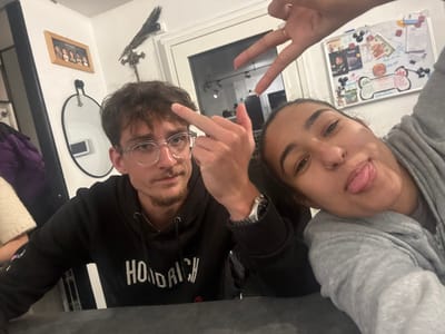 A man and a woman pose for a selfie. The man, wearing glasses and a black hoodie, holds up his middle finger. The woman, in a gray hoodie, sticks out her tongue and makes a peace sign with her hand. A picture frame and a whiteboard with drawings and writing are visible in the background.