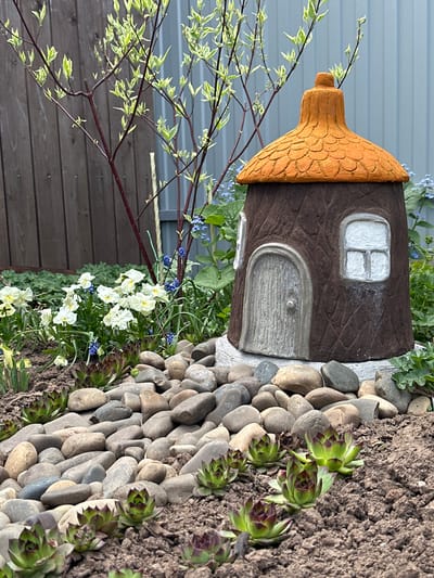 The image shows a miniature fairy house situated in a garden setting. The house is made of brown material with a textured wood-like appearance and has an orange, textured roof. A small, white-framed window and a gray arched door with a knob are visible on the house.
The house is surrounded by various plants, including white and blue flowering plants, and succulents. A border of gray river stones lines the edge of the garden bed. To the left of the house, a tree branch with small variegated leaves extends into the frame. A brown wooden fence is partially visible in the lower left corner of the image.
The location in Ludorvai, Russia, is not verifiable from the image.