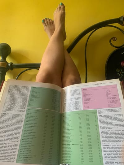 The image shows a close-up of a person's bare legs and feet, which are resting on an open book. The person's toenails are painted a vibrant turquoise. The legs are crossed and positioned over the book, suggesting a relaxed or contemplative posture. The book itself appears to be an atlas or encyclopedia, with pages filled with text and tables, some of which are visible in detail. The text is in Italian and includes entries for "Grenada" and "Grecia," along with statistical data and descriptive paragraphs about these locations. The background is a solid, warm yellow wall. The scene is lit with warm, indoor lighting, creating a cozy atmosphere. The overall impression is one of quiet study or leisure in a well-lit indoor space, possibly a study or living room. The mention of "San Quirico d'Orcia, Italy" in the prompt provides a specific geographical context for the scene, although it is not explicitly visible within the image itself.
